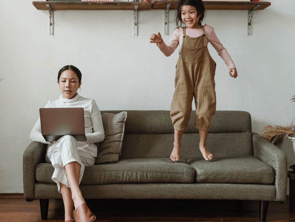 A mother working on the sofa with her daughter jumping next to her