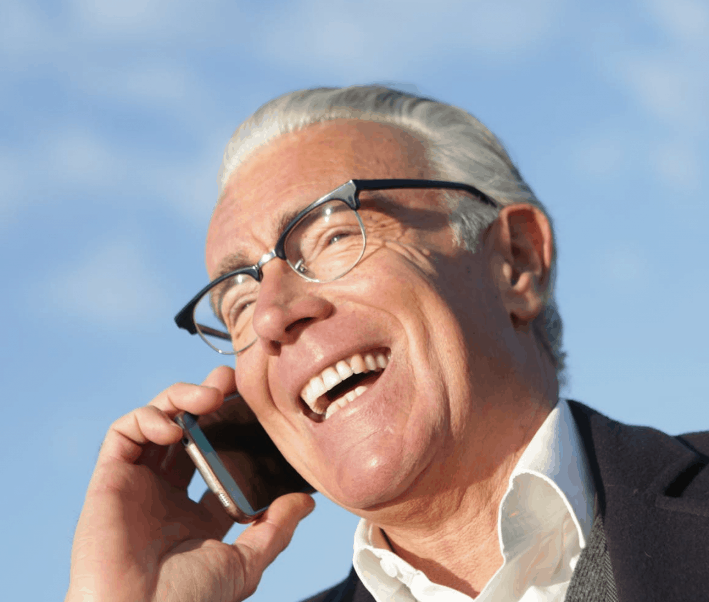 An elderly man smiling as he speaks on the phone outside 