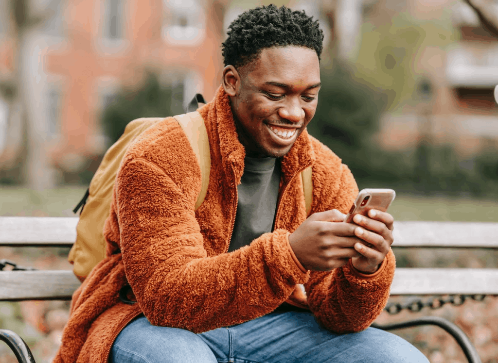 An African American man holding his phone and smiling on a bench